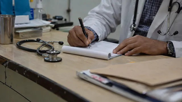 A doctor's hands writing a prescription on a pad at a consultation desk beside a stethoscope
